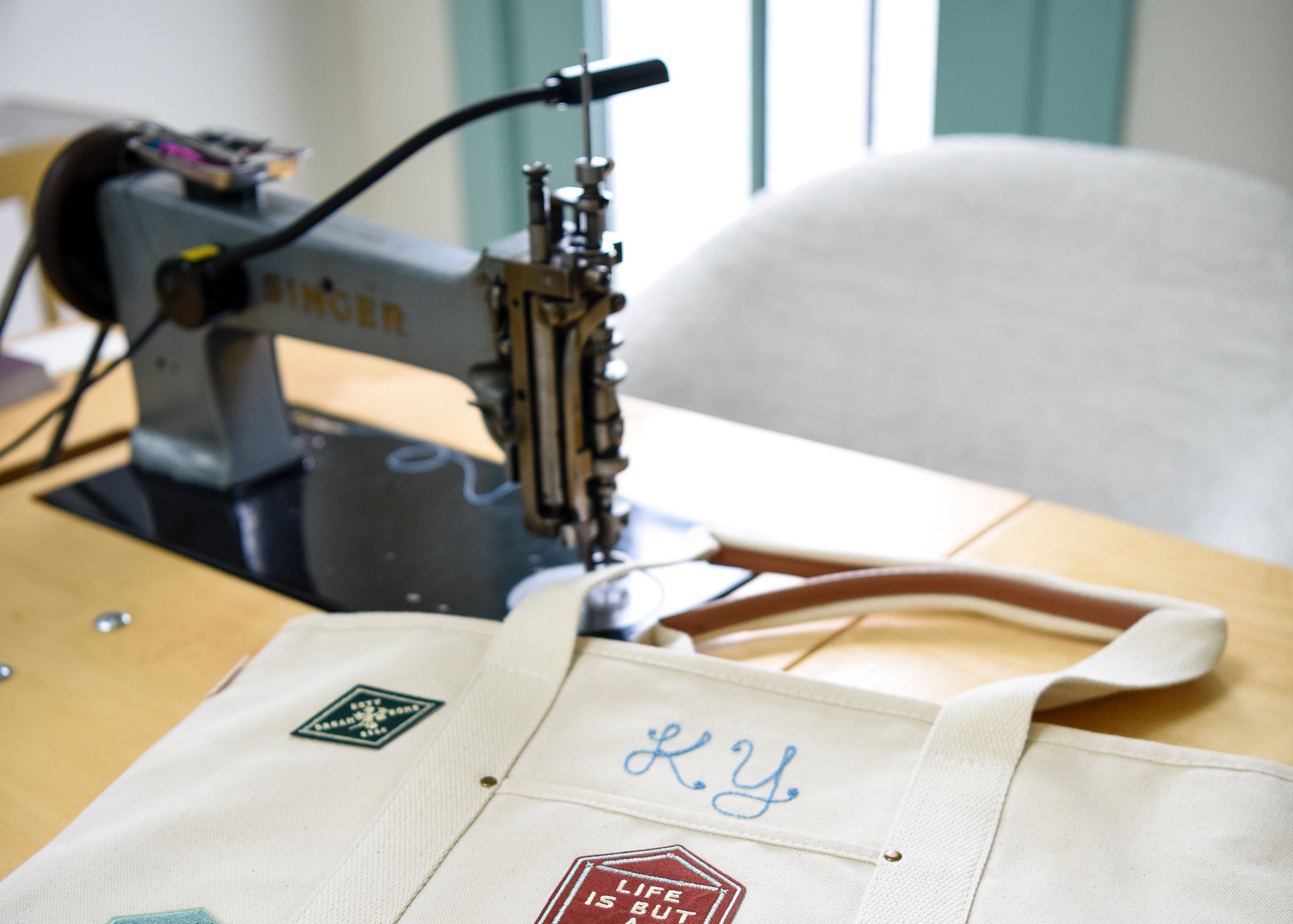 Customized white canvas tote bag with heat pressed patches on a wooden table with a chainstitch machine in the background.